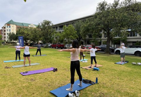 Group outdoor yoga session in a park with trees and buildings in the background.